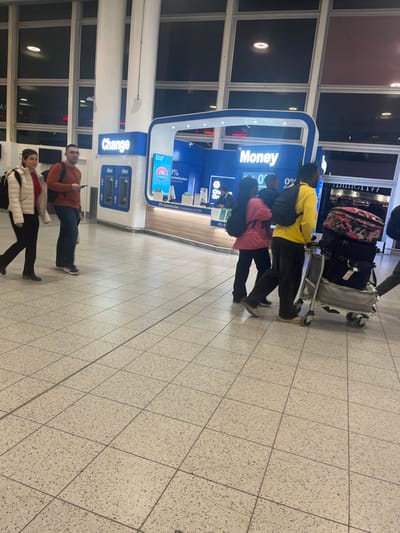The image captures an indoor scene at what appears to be an airport terminal, likely in Horley, United Kingdom (Gatwick Airport). The time of day is evening or night, suggested by the dark windows looking outside and the reliance on artificial lighting within the building.
In the mid-ground, a group of three people stands in front of a blue and white service point labeled "Money" in large white letters. One individual, wearing a bright yellow top, is positioned closer to the counter, pushing a metal luggage trolley laden with several bags, including one with a floral pattern. Behind them, another person in a pink or red top stands. The "Money" service point features screens displaying "0% Commission" and "City... Group," indicating it's likely a currency exchange or financial service provider. Adjacent to this main counter, a structure is labeled "Change" with "Atm" below it, revealing the presence of ATM machines.
In the foreground, two other individuals are either walking or standing. A man wearing an orange or brown long-sleeved top and dark trousers is visible, holding a mobile phone. Beside him, a woman in a white or cream puffer jacket and black trousers walks towards the right side of the frame.
The floor is covered with light-colored, square-tiled flooring. Large windows with dark reflections line the background, showcasing the indoor lighting and suggesting the exterior is dark. The overall atmosphere is functional and typical of a busy travel hub.
