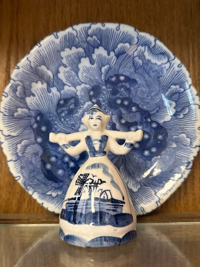 A ceramic figurine of a woman in traditional Dutch attire stands before a blue and white patterned plate. The figurine, painted with a windmill and other Dutch landscape elements, is positioned centrally in the frame. The plate, with its intricate floral or wave-like pattern, serves as a backdrop, filling most of the image. The items are displayed on a reflective surface, possibly a glass shelf, against a wooden background. The overall scene suggests a decorative display, likely within a home or a shop, in Graham, United States of America.