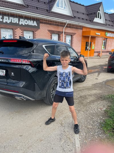 A young boy stands in front of a black SUV on a gravelly road. He is wearing a gray tank top with a wave graphic and dark shorts, and black athletic shoes. He is flexing his biceps, and his expression is determined. Behind him, a brick building with a white roof and brown trim houses a store called "For Home" (ДЛЯ ДОМА) and a "Monetka" (Монетка) grocery store. The building has orange accents and a green sign. The sky is partly cloudy. The image appears to have been taken in the city of Maminskoe, Russia.