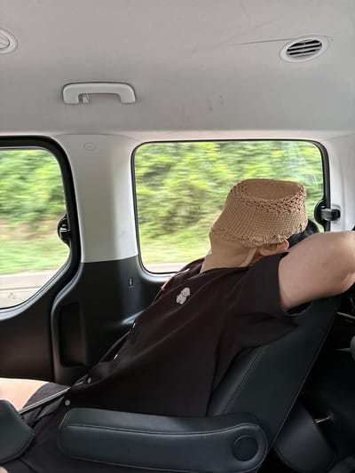 The image shows the interior of a vehicle, likely a minivan, with a person asleep in the back seat. The person is wearing a tan crochet hat and a dark t-shirt with a white graphic. They are leaning their head against their arm, which is resting on the seat. The vehicle's interior features gray headliner, air vents, a grab handle, and black leather seats. Through the windows, a blurred green landscape suggests the vehicle is in motion, possibly on a road in or near Seongpyeong-ri, South Korea, given the context provided by the user. The lighting is natural, coming from outside.