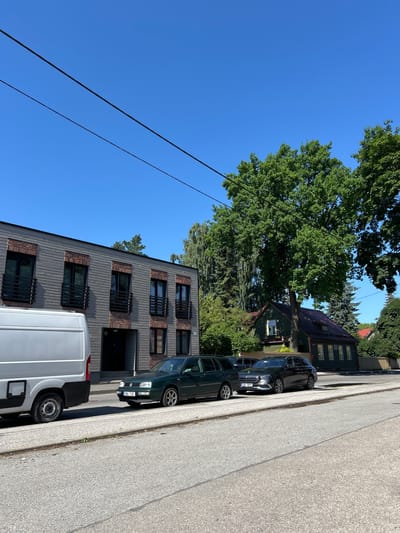 A low-angle, full shot captures a street scene in Tartu, Estonia on a clear, sunny day. The foreground is dominated by a grey asphalt road with visible cracks and a curb. To the left, the rear of a white delivery van is parked, its side facing the camera. Beside it, a dark green Volkswagen Golf station wagon is parked, facing right, with its license plate reading "504 TJC". Behind the Golf, a dark grey Mercedes-Benz E-Class station wagon is also parked, facing right, with its license plate reading "268 RPF".
The background features a two-story modern building with grey wood siding and dark-framed windows. Red brick accents are visible around some of the windows. A dark entrance is visible on the ground floor. To the right of the modern building, a traditional green house with a dark red roof stands under a large, mature oak tree. The house has several windows visible. Beyond the house, more trees and foliage create a dense green backdrop. Power lines stretch diagonally across the bright blue sky. No people are visible in the image.
