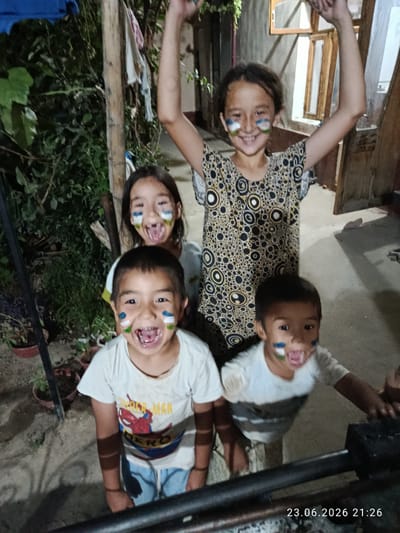 Four children, two boys and two girls, are in the foreground of the image, smiling and showing off face paint. The girl at the back, standing, has her arms raised. The other three children are in the front row, two boys and one girl. They all have what appears to be face paint resembling the flag of Uzbekistan on their cheeks. The girl in the front has face paint that also extends to her forehead and chin. The children are wearing casual clothing. The background shows an outdoor setting at night, with some vegetation, a wooden post, and a building with windows. The lighting is dim, suggesting it is evening. The overall mood is joyful and celebratory. Visible text in the lower right corner reads "23.06.2026 21:26".