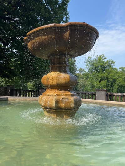 This is a full-frame, eye-level, vertical shot of a tiered stone fountain with water flowing and splashing. The fountain is made of light brown, rough-hewn stone. Water cascades from the upper basin, creating a curtain of droplets, and also splashes from the base into the surrounding pool. The pool water is a greenish hue and appears clear, with ripples and splashes visible. In the background, a wooden fence runs along a raised embankment. Beyond the fence are lush green trees and a bright blue sky with scattered white clouds. The scene is lit by natural daylight, suggesting it is daytime. The fountain is located in Náchod, Czechia.