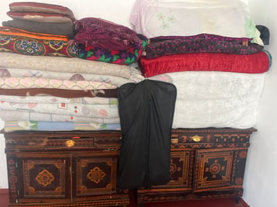 In a room in Tashlak, Kyrgyzstan, a collection of bedding is neatly stacked on top of an antique, dark wood dresser. The dresser features intricate inlaid patterns in shades of gold and orange. On the left side of the dresser, the bedding is stacked high, with visible layers including patterned blankets and what appears to be a comforter with a dark grey cover. Further to the right, a pile of folded blankets in various colors and patterns are stacked, including a bright red one and a purple one with fringed edges. In the center of the dresser, a black garment bag hangs down, obscuring some of the bedding. Behind the stacks of bedding, large white sheets are draped, one with a subtle floral pattern. The scene appears to be indoors, with a neutral-colored wall as the background. The lighting suggests it is daytime.