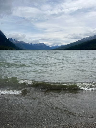 A scenic view presents a tranquil, gray-toned lake edged by a dark pebbly shore. Gentle waves lap the edge. Beyond the water, a mountain range climbs toward a cloudy sky, the peaks partly shrouded in mist. Patches of greenery cling to the mountainsides, contrasting with the gray and white tones of the scene.