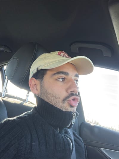 A male individual is depicted in the passenger seat of a vehicle. He has dark hair, stubble, and brown eyes, and wears a light-colored baseball cap with a red and white logo, along with a black ribbed turtleneck sweater featuring a silver half-zip. His head is turned towards the right, and his mouth is pursed into an 'o' shape. A dark-colored headrest is behind him, and a dark car door panel is to his right, with a grab handle visible on the ceiling. Bright daylight illuminates the scene from the right, through the vehicle's window, which shows an indistinct, blurred exterior background. The specific location within Mauléon-d'Armagnac, France, is not identifiable from the visual information presented.
