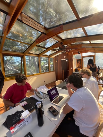 A group of people are gathered around a table inside a sunroom-like structure with large windows. Two are looking down at laptops, while a third person, wearing a red shirt, is reviewing papers. Items on the table include a water bottle, a phone, a catalog, and a package of Sharpie markers. Sunlight streams through the windows, illuminating the indoor space and highlighting the wooden beams and glass panes.
