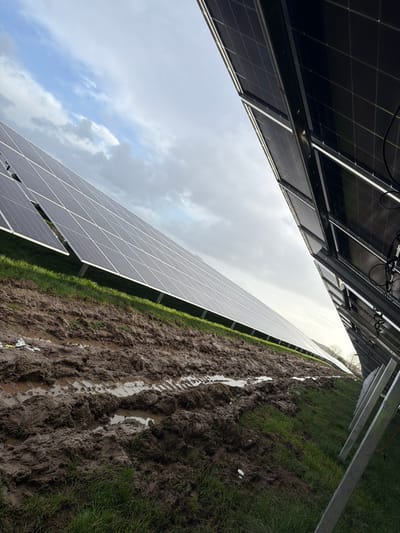 The scene depicts a solar farm located in The Municipal District of Adare — Rathkeale, Ireland. The foreground features a wide, deeply rutted path of dark brown mud, heavily saturated with water, displaying clear vehicle tire tracks. Several small pieces of white debris are visible within the mud on the left side. Beyond the muddy path, a strip of vibrant green grass extends to the base of multiple rows of dark, rectangular photovoltaic solar panels. These panels are mounted on light-colored metal support structures. The front, light-absorbing surfaces of a long row of panels are visible on the left, angled upwards. On the right, the undersides and associated wiring of another parallel row of solar panels are visible, with their support frames angled towards the viewer. The sky above is partially overcast, exhibiting a mix of grey and white clouds against patches of light blue, indicating a daytime setting with diffused lighting. No individuals are present or discernable within the frame.