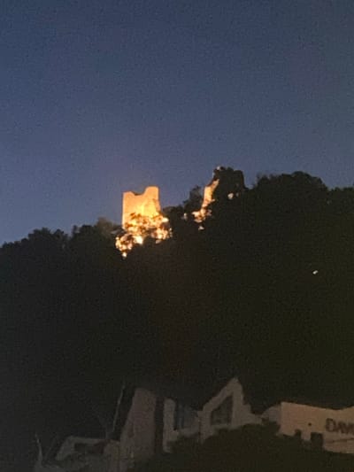 An image taken at night shows the ruins of a castle, likely Neamț Citadel, illuminated by warm lights from below. The castle is perched on a dark, tree-covered hill. In the foreground, the faint outlines of buildings and structures are visible, partially obscured by darkness and foliage. The sky is a deep, clear blue. The overall impression is of a historical landmark at dusk or after dark.