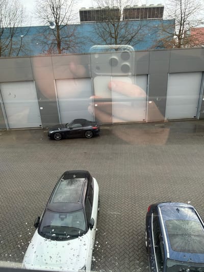 The image presents an exterior scene captured from an elevated position within Apeldoorn, Netherlands, looking down through a window visibly marked with numerous raindrops. A prominent reflection of a light-skinned hand holding a smartphone, equipped with a clear case and a three-lens camera array, is centered in the upper-mid portion of the frame.

The environment below consists of a paved surface made of gray interlocking bricks arranged in a herringbone pattern, appearing wet. In the background, a large building features a lower section paneled in gray, with multiple light-colored, closed roll-up garage doors. Above this, the building's upper section is painted light blue, topped by two visible industrial HVAC units. Several bare trees, devoid of foliage, are scattered between the paved area and the blue building section.

Three vehicles are parked on the wet brickwork:
1.  A black convertible sports car, featuring multi-spoke wheels and a partially deployed or retracted soft-top roof, is positioned in the mid-ground, angled towards the right in front of the gray building section.
2.  A white compact SUV or crossover, with a black roof and panoramic sunroof, occupies the left foreground, parked facing away from the building. Its roof surface is wet.
3.  A dark blue sedan or hatchback, also with a wet surface and visible sunroof, is partially visible on the right side of the foreground, similarly parked facing away from the building.