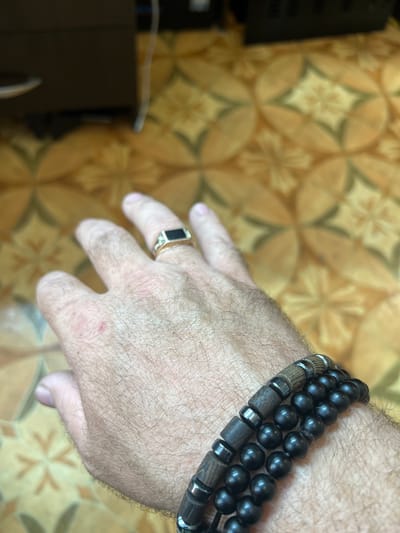 A close-up, high-angle shot captures an individual's right hand and lower forearm, set against a blurred, patterned floor. The hand, which appears to belong to an adult male given the visible hair on the back, is adorned with jewelry.

On the wrist, there are multiple stacked bracelets. One is composed of uniformly sized black spherical beads, while another features alternating dark, cylindrical wooden beads and smaller, shiny metallic ring-shaped spacers, possibly made of hematite or silver-toned metal. On the ring finger of the hand, a gold-colored ring with a prominent black, rectangular flat stone or inlay is visible. The skin on the back of the hand shows some minor reddish marks or imperfections.

The background consists of a floor covered with a repeating geometric pattern, likely tiles or linoleum. The pattern features interconnected light brown, petal-like shapes outlined in a darker shade, forming a symmetrical, star-like or floral motif at the center of each repetition. The background is significantly out of focus, emphasizing the hand and its accessories. In the lower-left corner, a dark, indistinct object, possibly furniture or electronics, is partially visible and heavily blurred. The lighting suggests an indoor setting, with no specific time of day discernible. No text is visible in the image. The location is Krayevsko-Armyanskoye, Russia.