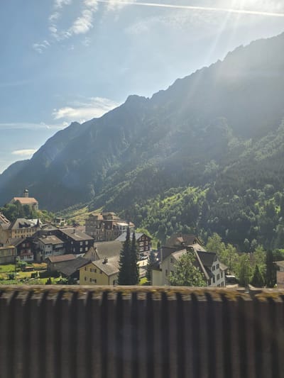 The image depicts a sun-drenched, mountainous village scene in Wassen, Switzerland, captured during the daytime. The foreground is dominated by a corrugated surface, possibly a roof or barrier, casting strong shadows. Beyond this, a picturesque village unfolds, with tightly packed houses featuring red-tiled roofs and light-colored facades. The architecture suggests a traditional European mountain settlement. The village is nestled at the foot of a steep, verdant mountain slope covered in dense forestation. Sunlight streams from the left, creating a bright haze and highlighting the contours of the mountain and the valley. Wispy white clouds are visible in the clear blue sky. The overall impression is one of tranquility and natural beauty, with the village harmoniously integrated into its mountainous environment. No people are distinctly visible, but the presence of houses implies inhabitants. The time of day appears to be morning or early afternoon, given the angle of the sun and the bright, clear conditions. There is no visible text in the image.