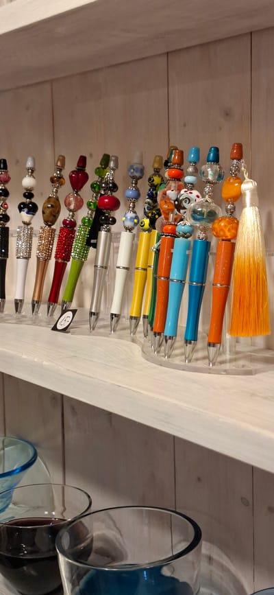 The image displays a collection of decorative pens with beaded tops, arranged on a white wooden shelf. The pens vary in color and design, with some featuring intricate beadwork and others adorned with large, colorful glass beads. A price tag reading "15-" is visible in front of one of the pens. The background consists of pale wooden planks. In the foreground, partially visible, are glass cups, one containing a dark liquid, possibly wine. The location is identified as Malda, Estonia.