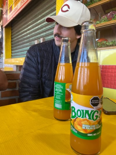 The image shows two glass bottles of Boing! mango juice, one larger than the other, lying on a yellow surface. The labels are prominently displayed, advertising "Bebida con 12% de pulpa de mango" and a "Exceso Calorias" warning. A person wearing a baseball cap and a dark jacket is partially visible beside the bottles. The background includes a blurred scene with a sign that says "Bienvenidos!" and a glimpse of a building exterior.