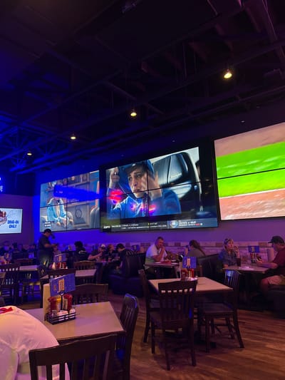 The image shows the interior of a sports bar in College Station, Texas, United States. Several large screens display sports games, including basketball and baseball. Multiple tables are arranged in rows, with people seated at them, eating and watching the games. Condiments and menus are visible on the tables. The lighting is a mix of ambient and colored, with blue and yellow hues prominent. Exposed ceiling beams are also visible.