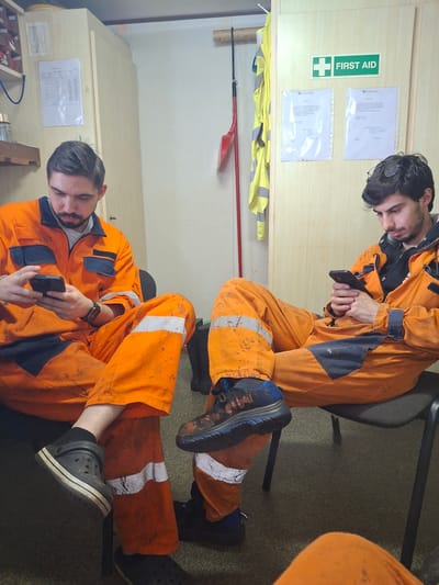 Two individuals wearing orange work jumpsuits are seated indoors. The man in the foreground is wearing black Crocs and looking at his phone. The second man in the background is wearing dark sneakers and is also looking at his phone. Behind them is a wall with a "First Aid" sign, documents, and a yellow vest hanging. The specific location in Cuxhaven, Germany cannot be determined from the image.