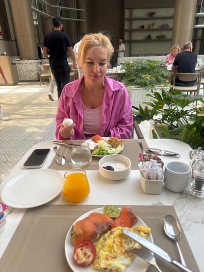 A woman sits at a table with a partially eaten breakfast in El Gouna, Egypt.
The woman is blonde and wearing a pink shirt and a white tank top. She is holding a white egg in her left hand.
On the table, there is a plate with salmon, eggs, and a tomato, a bowl with yogurt and olives, a salad bowl, a glass of orange juice, a glass of water, a knife and fork, a spoon, an empty plate, a smartphone, and a container of sugar packets. There are salt and pepper shakers and a coffee cup on the table as well.
In the background, there are other people sitting at tables. There are plants and buildings visible. The floor has a geometric pattern.