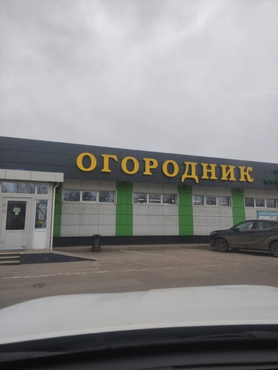 The image captures the exterior of a modern commercial building under an overcast sky. The building, positioned on the left, features a contemporary design with dark gray panels and alternating horizontal stripes of green and white. Large yellow Cyrillic letters prominently display "ОГОРОДНИК" (OGORODNIK), which translates to "Gardener" or "Market Garden," on the dark gray facade, indicating the nature of the business.
Below the main signage, the building has several windows, some reflecting the cloudy sky and bare tree branches. A glass entrance door is visible at the bottom, and a black trash can stands nearby.
Further text details are visible on the building: a sign on the green stripe near a parked car reads "СЕМЕНА САЖЕНЦЫ" (SEMENA SAZHENTSY), meaning "Seeds Seedlings." Below this, phone numbers are listed: "+7-999-854-21-54" and "+7-999-112-27-75". Inside one of the windows, the words "ИНТЕРНЕТ МАГАЗИН" (INTERNET MAGAZINE) are discernible, suggesting an online store component.
To the right of the building, a paved area, likely a street or parking lot, is visible. A dark-colored SUV or crossover is parked facing away from the viewer, with only its rear visible. Part of a white vehicle is also seen in the extreme foreground on the right. The sky is uniformly gray, suggesting a cloudy day. No people are visible in the scene. The Cyrillic text confirms the location context of Vereya, Russia.