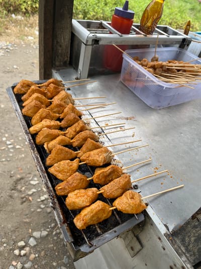 This image captures an outdoor street food stall in Matangpanyang, Indonesia, displaying various skewered items for sale. The primary focus is a rectangular metal charcoal grill rack on the left, holding 25 wooden skewers arranged in five rows of five. Each skewer holds one or more pieces of light orange-brown, irregularly shaped, grilled food, consistent with fried tofu (tahu goreng) or a similar fried snack, coated in a reddish seasoning or sauce. The grill rack itself shows signs of heavy use, with black charring and some remnants of drippings.
To the right of the grill, on a metallic countertop, a clear rectangular plastic container holds a quantity of smaller, roundish, light-colored skewered items, consistent with meatballs or fish balls (bakso), also on wooden skewers. These items are not currently on the grill but are prepared for cooking or serving.
Behind the container, a metal rack holds two bottles: a red plastic squeeze bottle with a purple cap, typically used for chili sauce or ketchup, and an inverted, clear brown bottle, likely containing soy sauce or another liquid condiment.
The stall appears to be a basic, mobile setup, with a dark wooden vertical support beam visible on the left. The ground immediately surrounding the stall is unpaved, consisting of dirt mixed with small grey and white pebbles and rocks. In the background, beyond the stall, dense green foliage is visible under natural light, indicating an outdoor, possibly semi-rural or roadside, setting. No individuals are visible in the frame.