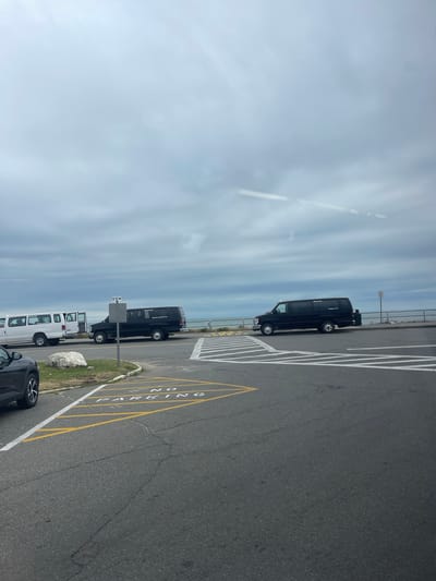 The image shows a row of parked vehicles, including black and white vans, on an asphalt surface marked with white and yellow lines. The vans appear to be in a parking lot. One black van has text on its side that reads "OCEAN VIEW TRANSPORT". Another white van is partially visible, with its doors open. In the foreground, a black car is parked. The background features a railing and a hazy sky with some clouds. The scene suggests a parking area, possibly near a waterfront given the presence of the railing and the expansive sky. The lighting indicates it is daytime, with a soft, diffused light. There is a signpost visible near the black van. The overall impression is of a calm, overcast day in a parking area. Visible text includes "OCEAN VIEW TRANSPORT" and "NO PARKING" stenciled on the asphalt in yellow.