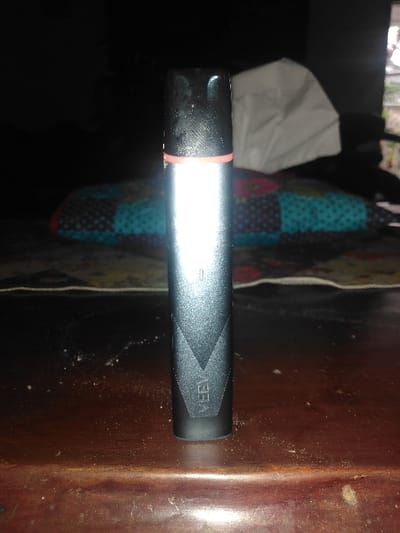 The image displays a dark-colored, cylindrical VEEV branded electronic vaping device standing vertically on a dark, polished wooden surface. The device features a matte black body, a glossy black mouthpiece/cap, and a thin red band positioned below the cap. A prominent white reflection illuminates the upper half of the device. The lower body of the device bears a textured pattern incorporating the vertically oriented brand name "VEEV." The wooden surface shows traces of dust and minor scuff marks. The background is dimly lit and out of focus. Immediately behind the device, a multi-colored patterned textile object, resembling a cushion or blanket with teal and dark blue polka-dotted square patches, is visible. To the right of the device, a crumpled white object, consistent with a tissue box or a pile of tissues, is present. A faint, blurred view of what appears to be outdoor greenery is discernible through a distant opening or window in the far background. The scene is static, devoid of people, and appears to be an indoor setting in Uccle - Ukkel, Belgium.