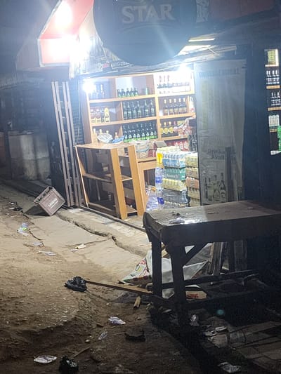 The image shows a street scene at night in Obiaruku, Nigeria. The location appears to be a small commercial area. A store is visible with shelves stocked with drinks, including bottled water and alcoholic beverages. A banner reading "UNIVERSE GLOBAL" and "Drinks Avail..." hangs above the store. A "STAR" sign is visible on the left. The ground is littered with debris and trash. A wooden table and various metal structures are present in the foreground, partially obstructing the view. A crate labeled "LEGEND" is lying on the ground. No people are clearly visible. The lighting is uneven, with bright areas near the store and dark shadows elsewhere.