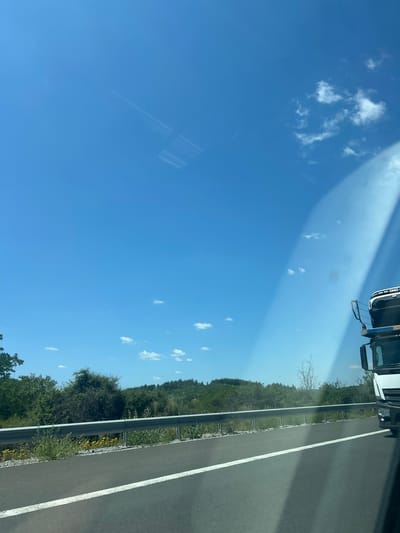 A truck drives on a highway on a sunny day. The truck is white with black trim. The highway has a white line down the middle. To the left of the highway is a guardrail, and beyond that is a grassy area with some yellow flowers. Further back is a dense line of trees and hills. The sky is bright blue with a few scattered white clouds. There is a glare on the windshield of the vehicle from which the photo was taken. The location is the highway near Rajana, Bulgaria.