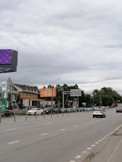 The image depicts a cloudy day scene on a multi-lane road in Trubacheyevka, Russia. The road is lined with guardrails and features white lane markings. In the foreground, traffic is moving in both directions. On the right side of the road, a white SUV with an orange stripe on its rear is visible, followed by a dark sedan. Further down the road, a convoy of vehicles, including a truck and several cars, is moving slowly.
On the left side of the road, a large billboard with a checkered purple pattern is mounted on a tall structure. Adjacent to it is a gas station with a blue and white logo. Further back, a row of houses with distinctive architecture, including dark wood siding and multiple gabled roofs, is present. A bright orange billboard with Russian text is also visible above the houses, advertising a travel company. Trees and vegetation border the road and the buildings. The overall impression is of a busy suburban or semi-urban environment.