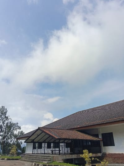 A building with a dark tiled roof fills the lower right of the frame against a bright sky with scattered white clouds. The building is white with dark-framed windows. An open porch with a smaller tiled roof extends from the building, supported by white columns. Trees are visible to the left, and paved ground extends around the building. The image captures a clear, sunny day with the building prominently featured.