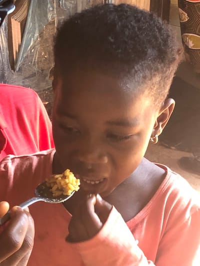 A young girl with dark, curly hair eats from a spoon. She is angled to the left of the frame and wears a pink long-sleeved shirt. Her eyes are partially closed, and she smiles, showing her teeth as she brings food to her mouth. The spoon holds rice or grains of a yellow-brown color. A hand partially visible in the foreground appears to be holding the spoon. The background includes blurred items, possibly furniture and clothing, in shades of black, brown, and beige.