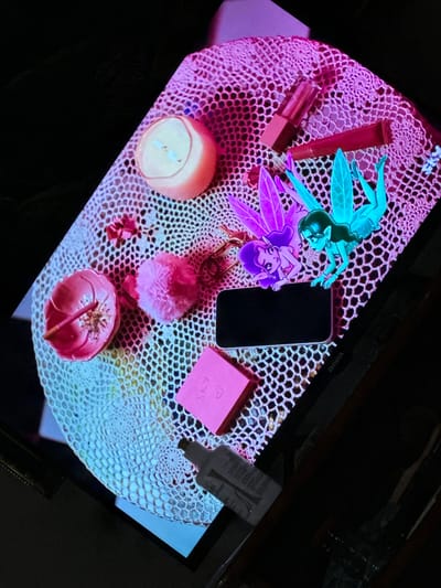 A still-life composition sits on a crocheted, oval-shaped surface, likely a doily, with a hexagonal mesh pattern. The items on the doily include: two lip gloss tubes, one lying horizontally and the other vertically; two cartoon fairy figures, one light blue and one purple; a dark, rectangular mobile phone; a pink square object with "SKY" written on it; a grey bottle with white lettering; a small, round candle in a pink holder; a small, pink pom-pom; and a pink flower-shaped dish containing a brown object. The doily is positioned on a dark surface, possibly a table. The image is bathed in a mix of warm and cool light, creating a purple hue on one side and a cooler, bluer hue on the other. The composition appears to be situated within a residential indoor environment. There is no indication this is in the city of Parroquia Ocumare, Venezuela.
