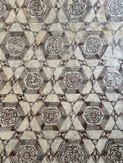 The image shows a patterned floor in Tepoztlán, Mexico. The floor is composed of repeating geometric shapes - hexagons containing floral designs and equilateral triangles. The designs are primarily in shades of brown and white, with a worn and aged appearance.