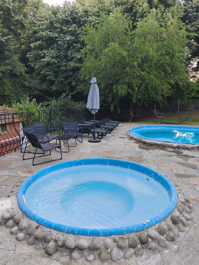 In the city of Gramatikovo, Bulgaria, a serene outdoor scene unfolds. Two circular blue pools, one larger and closer to the viewer, and another smaller one positioned further back, are the central focus. The pools are partially surrounded by an array of large, rounded stones, creating a naturalistic border. The ground surrounding the pools is paved with irregular, flat stones, forming a rustic patio.

To the left of the foreground pool, a black metal fence with intricate scrollwork stands, separating the patio from a brick structure with a window. Beyond the fence, lush green trees and foliage create a dense, verdant backdrop, with glimpses of the sky peeking through the leaves. A gray umbrella, closed and resting on its base, stands near a table with a glass top and four black chairs, all made of mesh material. Further back, several black lounge chairs are arranged in a row.

In the distance, more trees and vegetation can be seen, with hints of recreational equipment like a red swing and a green seesaw-like structure visible among the greenery. The overall impression is one of a peaceful, private outdoor space designed for relaxation and enjoyment.