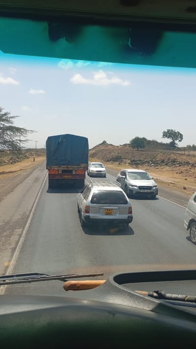 This image, captured from within a vehicle, presents a forward view of a multi-lane asphalt road in Malewa West ward, Kenya, under a clear sky with scattered white clouds. The immediate foreground shows the dark gray dashboard of the originating vehicle, upon which a light-colored wooden stick or club and another object with a dark, possibly wrapped, handle are resting. The upper section of the windshield displays tinting and some localized imperfections.
Traffic is observed on the road. Directly ahead, a large truck with its cargo covered by a blue tarp occupies the left lane. Its rear features red and yellow reflective tape and a license plate partially visible as "T90L 9797". To the right and slightly behind the truck, a silver Toyota station wagon, bearing Kenyan license plate "KAZ 108K" and equipped with a roof rack, is positioned in the adjacent lane. Further ahead in the right lane, a white minivan or small SUV is visible. In an opposing or adjacent lane to the right, a silver Subaru SUV with Kenyan license plate "KDS 914Y" is present. The front-right section of another white vehicle is partially visible on the far right.
The surrounding environment depicts an arid to semi-arid landscape. To the left of the road, a dirt shoulder is visible, beyond which sparse, thorny trees and dry vegetation grow. A single individual, dressed in dark clothing, stands on this dirt shoulder in the mid-distance. The background consists of rocky, brown hills and outcrops, dotted with a few isolated trees, under bright sunlight.