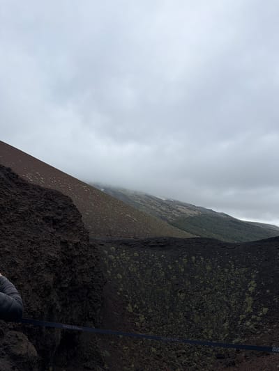 The image captures a landscape near Nicolosi, Italy. A dark, rocky terrain dominates the foreground, transitioning to a slope covered in sparse vegetation. A dark strap extends from the top right corner down the side. A blanket of clouds obscures the distant scenery.