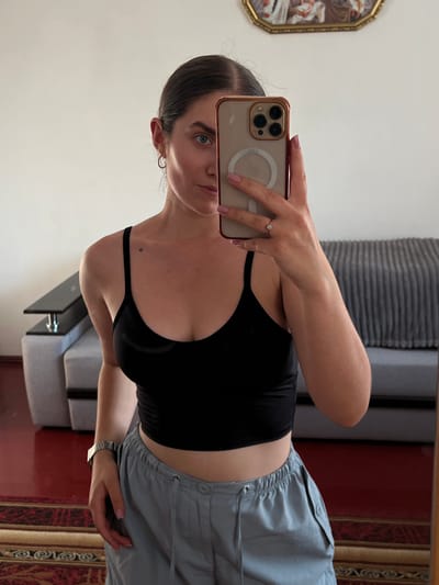 This media file captures a woman taking a mirror selfie. She is wearing a black crop top and light blue drawstring pants. A ring is visible on her left hand, and a watch on her right wrist. In the background, a piece of artwork in an ornate gold frame hangs on a white wall. To the right of the artwork, a grey, ribbed surface is visible, possibly a headboard or furniture. Further in the background, a sofa is partially visible, and the scene appears to be indoors, likely in a domestic setting. The lighting suggests daytime. There is no visible text in the image.