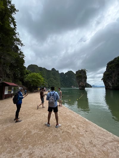 This image depicts a scenic coastal area with distinctive limestone karst formations rising from the water. In the foreground, a sandy path meets the water's edge, with several people present. One person in the foreground is wearing a blue shirt and black shorts, with a checkered backpack. To their left, another person is wearing a blue jacket and black leggings, holding a phone. Further back, more individuals are visible, some walking and others standing. The environment features lush green vegetation covering the rocky islands, and the water is a murky green. The sky is overcast with heavy clouds. The scene is identifiable as Khao Phing Kan, also known as James Bond Island, located in Phang Nga Bay, near Tambon Krasom, Thailand, famous for its unique geological formations and its appearance in the James Bond film "The Man with the Golden Gun."
