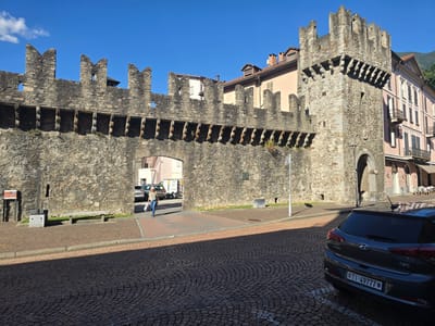 The image depicts an outdoor scene in Bellinzona, Switzerland, under a clear blue sky, suggesting a sunny day. The primary subject is a medieval stone wall with crenellations and an arched gateway. In the foreground, a dark grey car is parked on a cobblestone street, with its license plate reading "TI-49777".
A woman wearing jeans and a light-colored top is walking away from the viewer, carrying a white shopping bag, through the arched gateway. Beyond the gateway, cars are visible, and the entrance to a building is suggested. To the right of the wall, a multi-story building with balconies and windows painted in a soft pink hue stands adjacent to the ancient structure. Further in the background, more buildings and distant hills can be seen.
A stone bench is placed against the wall in front of the gateway. A signpost is also visible near the woman. The sun casts long shadows across the cobblestone street, indicating it is likely late morning or early afternoon. The overall atmosphere is one of historical charm combined with everyday life.
