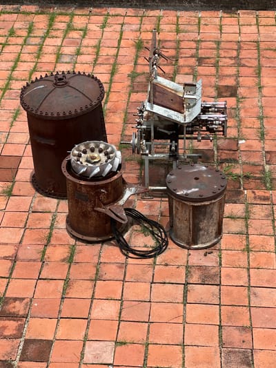 In the city of San Agustín Etla, Mexico, four rusty metal cylindrical objects are arranged on an outdoor paved area made of reddish-orange bricks with grass growing between them. To the left is a tall, wide cylinder with a domed, bolted top. Next to it, and slightly forward, is a shorter, narrower cylinder with a fan-like object inside. To the right of the first cylinder is a smaller, squat cylinder with a flat, bolted top. Behind these objects is a complex, multi-component metal apparatus with gears, rollers, and wooden accents, mounted on a metal frame. A black coiled cable lies on the ground near the shorter, narrower cylinder. The scene is viewed from above.