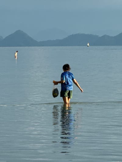 The image depicts a serene coastal scene at Baan Pang Ka, Thailand. In the foreground, a person stands in shallow water, facing away from the camera. They are wearing a blue t-shirt and green shorts, and are carrying a clear plastic bag filled with dark objects. The water is calm with ripples reflecting the sky and the distant landscape. Further in the distance, another person is visible wading in the water. On the horizon, a small sailboat can be seen, and behind it, layers of hazy mountains form the backdrop. The overall atmosphere is tranquil and serene, with the muted colors suggesting either early morning or late afternoon light.