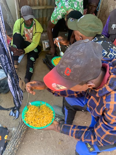This media file shows a group of people eating together outdoors. The subjects are several individuals, some wearing caps and casual clothing, gathered around to share a meal. One person is holding a plate of noodles and beans with a spoon, in the process of eating. Another person in a green jacket and cap is seated, with their hands clasped. The scene appears to be a casual outdoor gathering, possibly in a rural or less developed area, given the ground surface which is a mix of dirt and gravel, and the presence of what looks like a wooden log.

The activity is clearly eating, suggesting a communal meal or a break for food. The clothing of the individuals indicates a warm climate, likely during the day. One person is wearing a blue and orange plaid shirt, another a green long-sleeved shirt and a grey cap, and another a green shirt with a white pattern.

Visible text includes "RTCAP" on a green cap, and several instances of "Calvin Klein" on a black and white ribbon or fabric strip lying on the ground. There are also some indecipherable characters in what appear to be Asian script on a black bag in the background. The word "Sup" is visible on a yellow and black tag on a blue garment.

The overall impression is one of a simple, communal meal shared in an informal setting. There are no strong cues about emotions, but the act of sharing food often implies togetherness and a sense of community. The location context provided places the scene in Boi A, Nigeria.