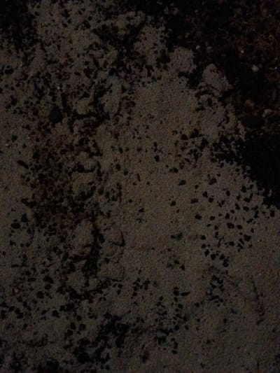 The image shows dark, textured ground or soil, possibly a mix of sand and small pebbles. The lighting is dim, casting shadows that emphasize the uneven surface. There are subtle variations in color and texture, suggesting a heterogeneous composition of the ground. The darker areas might indicate moisture or denser material, while lighter parts suggest drier sand.