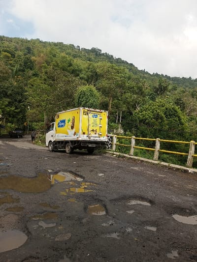 A yellow ice cream truck is parked on a pothole-ridden road. The truck has a white cab and displays an ice cream cone graphic. A railing painted with yellow and white stripes borders the right side of the road. A person walks along the road in the distance. A lush green, tree-covered hill rises in the background under a cloudy sky. Reflections of the truck are visible in the water-filled potholes.