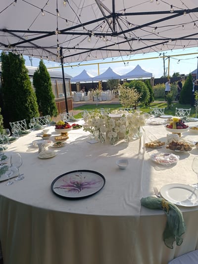 The image depicts an outdoor event setup in Pchegatlukay, Russia, under a large white tent adorned with string lights. A long, round table is set for a meal, draped in a cream-colored tablecloth. Place settings include white plates, clear wine glasses, and silverware, with some tables also featuring a decorative plate with a floral design. Centerpieces of white flowers in glass vases are arranged on the table, along with bowls of fruit, pastries, and other food items. In the background, more tents are visible, as well as people mingling and seated at other tables. The setting appears to be a festive occasion, possibly a wedding or celebration, with lush green landscaping surrounding the area.