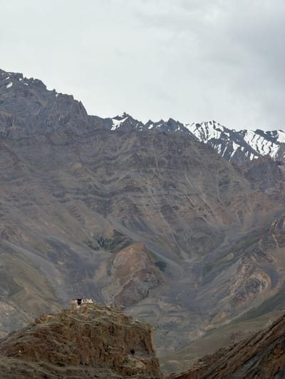A remote monastery sits atop a rocky outcrop in Zangla, India, under a cloudy sky. The monastery, with its white walls and red roof, is small and appears to be of traditional architecture. It is perched on a steep, rugged hill, surrounded by barren, mountainous terrain. The mountains in the background are imposing, with jagged peaks and slopes covered in scree and rocks. Some of the higher peaks are dusted with snow. The overall impression is one of isolation and grandeur, typical of the high-altitude regions of India.
