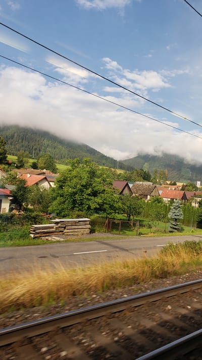 The image depicts a rural village scene in Kráľova Lehota, Slovakia, viewed from a moving train. In the foreground, train tracks run horizontally across the bottom of the frame. Beyond the tracks, a strip of dry, golden grass borders a paved road with a white line marking its center. To the left of the road, a stack of lumber is piled. Further back, a cluster of houses with red-tiled roofs and some with grey shingles are visible amidst lush green trees and manicured lawns. The village is nestled at the foot of a densely forested mountain range, which is partially obscured by low-hanging clouds and mist. The sky above is a clear blue with scattered white clouds, and overhead power lines stretch across the upper portion of the image.