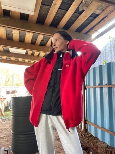 A person with dark hair and light skin is wearing a red jacket over a black jacket and light grey sweatpants. They are standing next to a black barrel. The background consists of wooden planks, a corrugated metal structure, and an outdoor area with vegetation. The location is Doma 45 Kilometr, Russia.