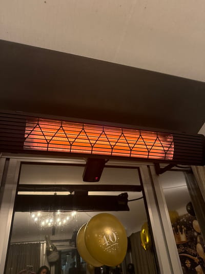 A black, vertical space heater with a glowing orange interior stands next to a window. The window reflects a party scene with gold balloons, one of which has "40" printed on it. The heater is mounted on the wall and appears to be in operation, radiating heat. The background suggests a celebratory event is taking place.