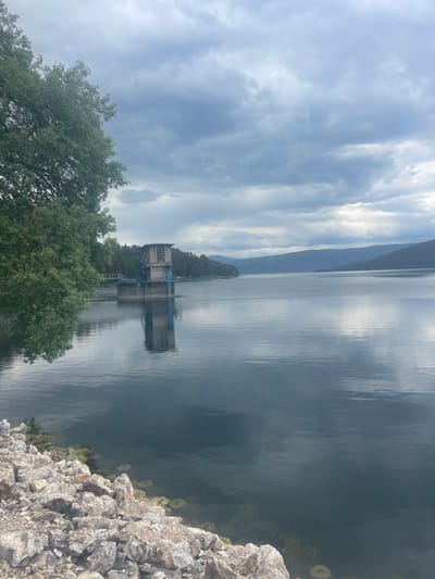 The image shows a building extending into a body of water, likely a lake or reservoir, with a mountainous terrain visible in the distance. The sky is overcast with a mix of light and dark clouds. In the foreground, there are trees and a rocky shoreline. The building appears to be a water control or pumping station, with pipes and a structure partially submerged. The water's surface reflects the sky and the building. Given the city context, this is likely situated around the area of Sv. Konstantin, Bulgaria.