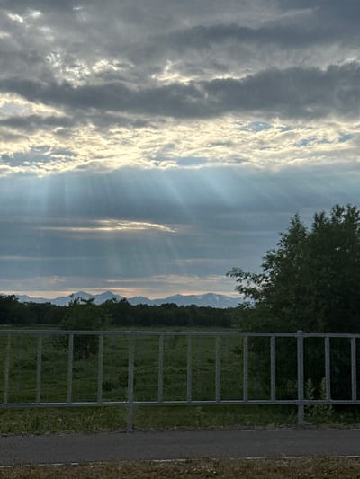 The image depicts a scenic landscape with a cloudy sky casting rays of light. A metallic fence runs horizontally across the foreground, separating a paved path from a grassy area. In the background, a dense line of trees stretches across the horizon, with mountains visible in the far distance. The overall scene is one of natural beauty and tranquility. It is impossible to determine the specific location of Кеткино, Russia from the provided image.